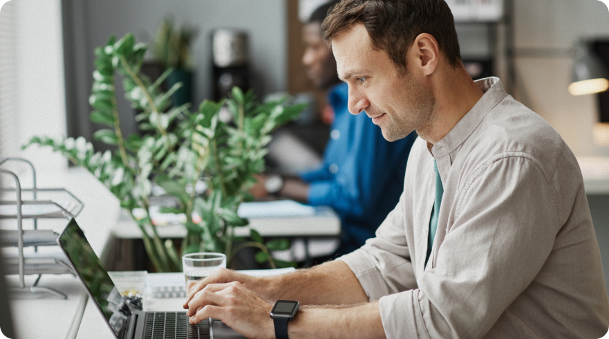 A side profile shot of a man in a light-colored shirt, sitting at a desk and typing on a laptop. He is wearing a smartwatch, and a glass of water and a potted plant are visible on the desk. This image represents the concept of **continuous endpoint monitoring** and highlights the use of a laptop in a modern office environment.