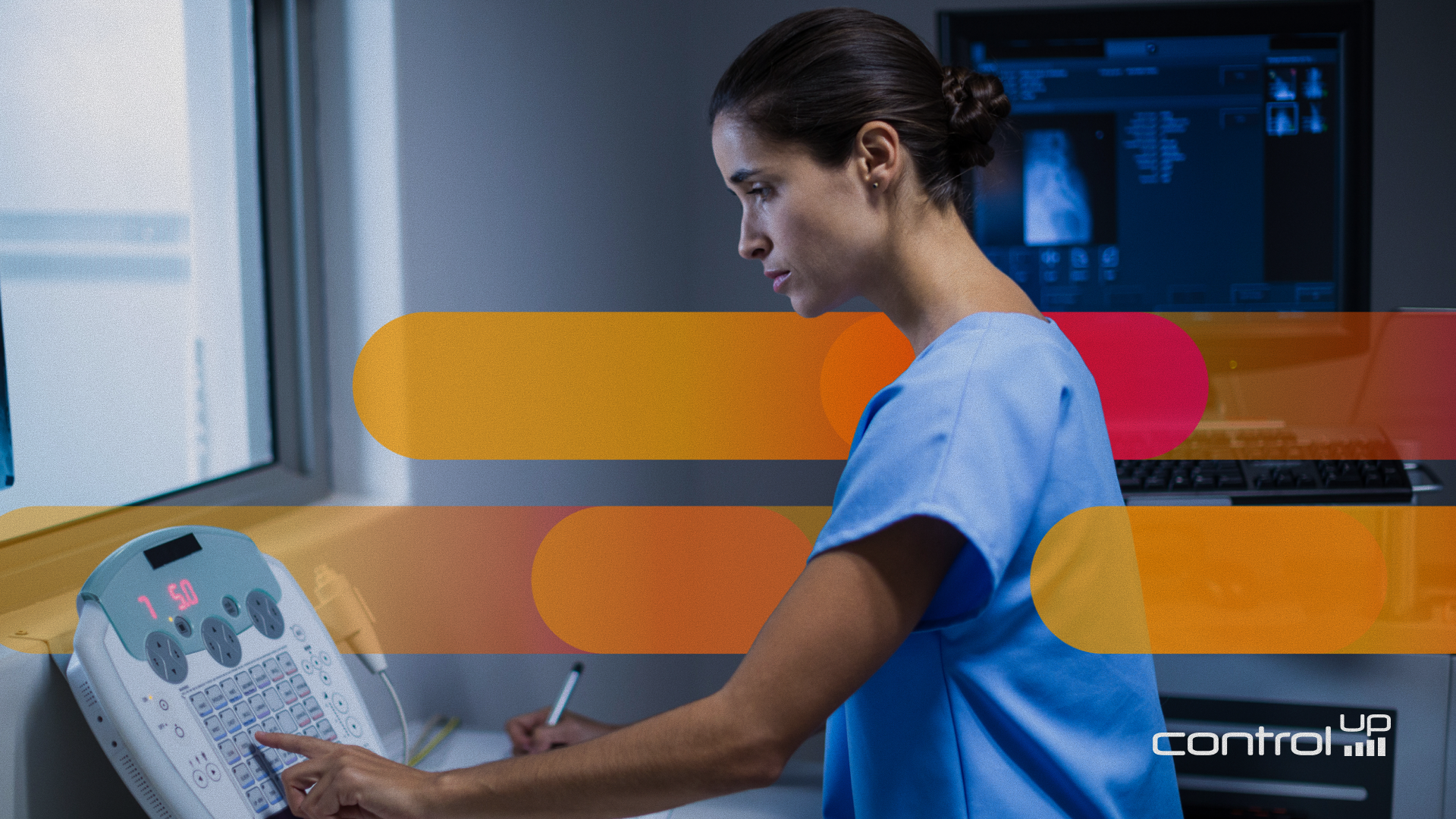 female with dark brown hair pulled into a bun and blue scrubs looking down at a healthcare machine in a dimly lit hallway