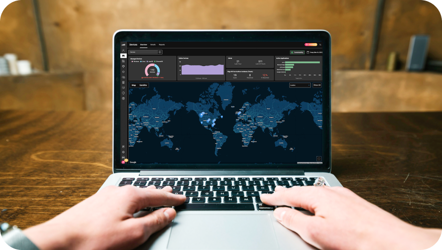 A first-person perspective of hands typing on a laptop placed on a wooden desk. The screen displays a dark-themed data analytics dashboard featuring a world map with highlighted locations, along with various graphs and performance metrics.