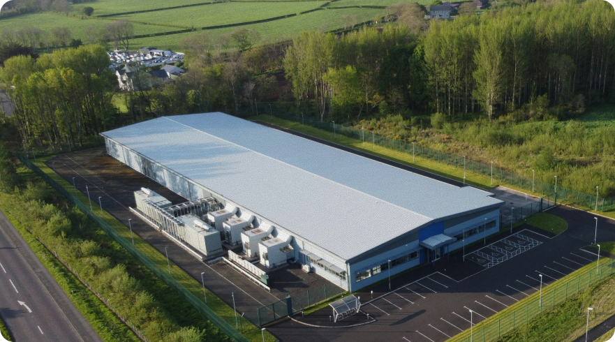 Aerial view of a long, single-story data center building with a gray metal roof, surrounded by green trees and grass. The building has a large, paved parking lot and is situated next to a road. The image is used to illustrate IT sustainability and Scope 2 emissions from IT data center infrastructure.